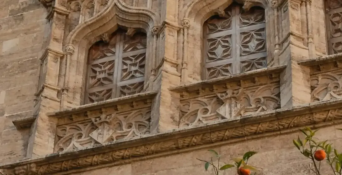 Edificio de piedra ornamentado con ventanas góticas, detalles tallados y una bandera, parcialmente cubierto por un naranjo co