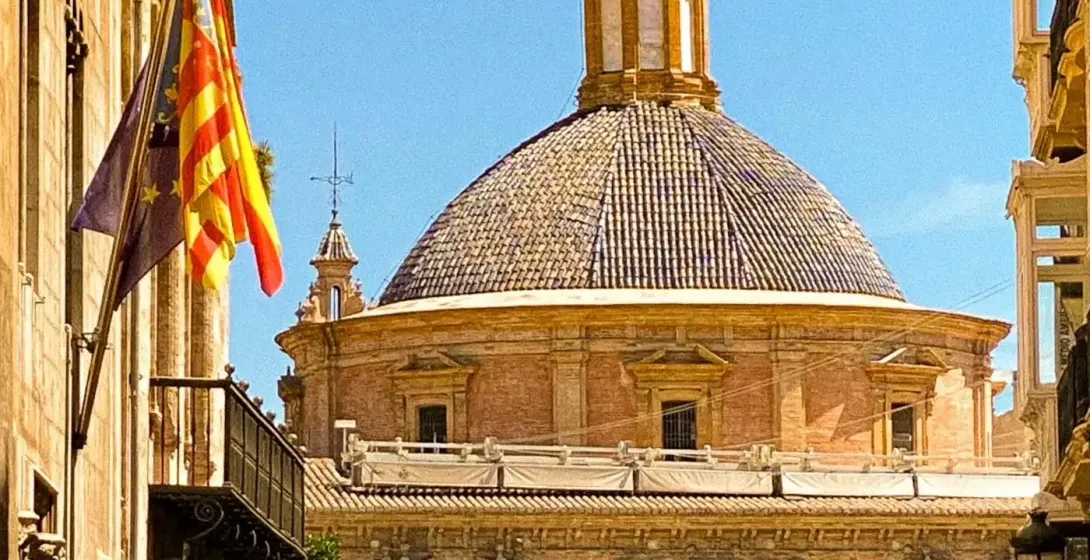 Estrecha calle peatonal en Valencia con gente caminando, que lleva a un edificio religioso con cúpula de tejas y linterna baj