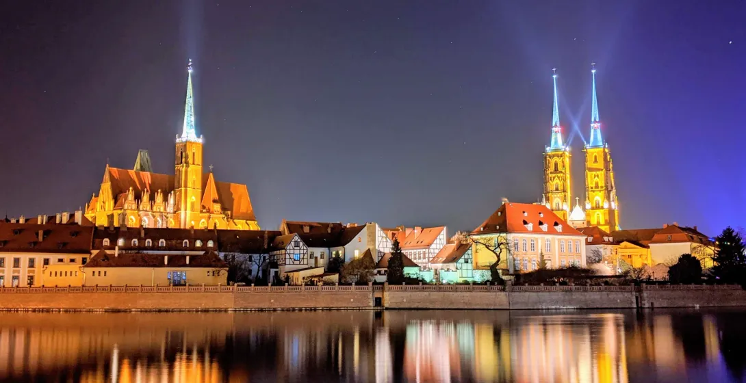 Wroclaw's Cathedral Island (Ostrów Tumski) at night, with illuminated historic churches and buildings reflected in the Odra R