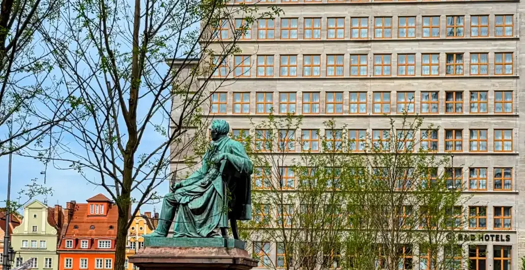 Bronze statue of a seated man on a plinth in a city square, with buildings and trees in the background.