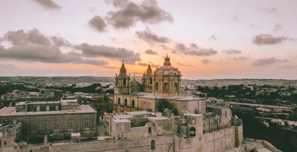 Aerial view of St. Paul's Cathedral in Mdina, Malta, with a sunset sky and historic city walls.
