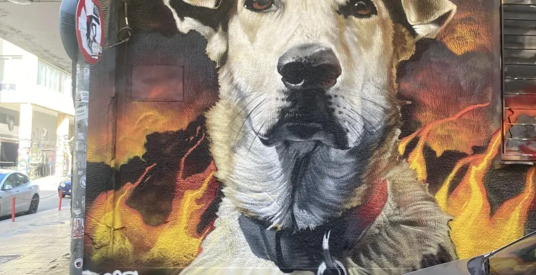 Large street art mural of a light-furred dog with a golden crown and flames in the background, on an Athens street.