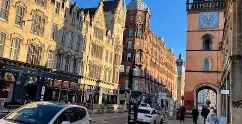 Historic street in Glasgow with ornate stone buildings, a prominent reddish-orange clock tower, and a white car parked on ...