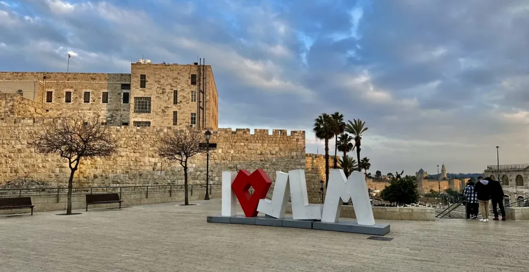 Touristen besuchen das ikonische "I ♥ JLM"-Schild in Jerusalem, mit den alten Stadtmauern im Hintergrund.