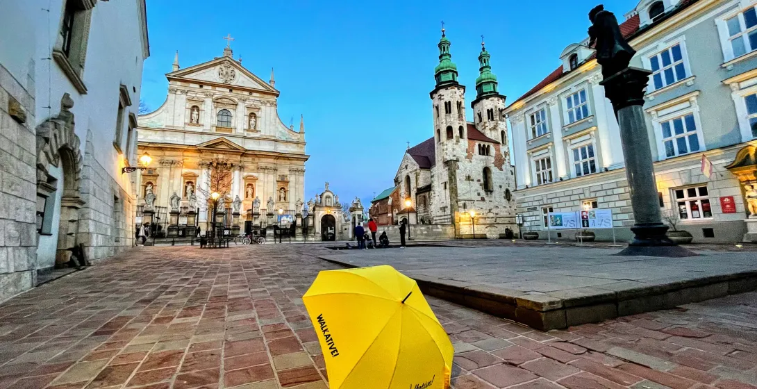 A yellow umbrella with 'Walkative!' in Krakow's Old Town square at dusk.