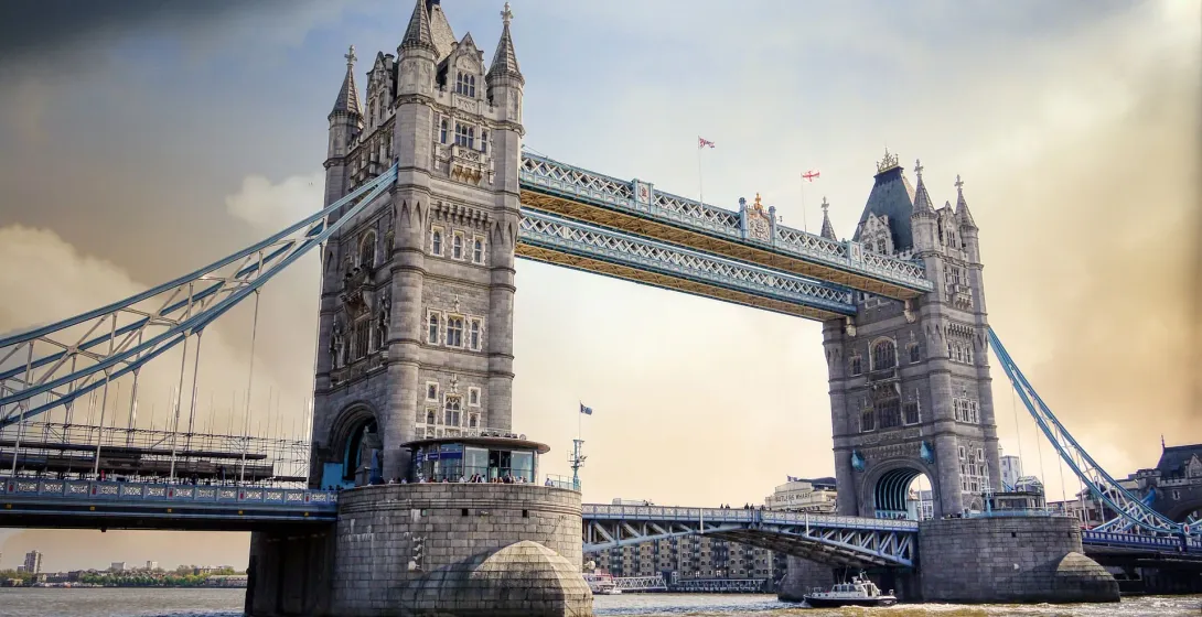 Most Tower Bridge rozciągający się nad szeroką rzeką pod częściowo zachmurzonym niebem, z łodzią na wodzie i budynkami w tle.