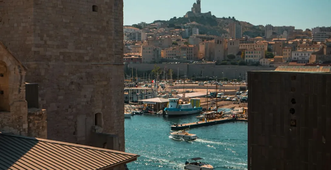 View from a stone fortress over Marseille's Old Port with boats and Notre-Dame de la Garde basilica on a hill.
