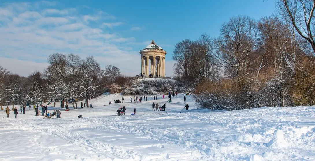 Ośnieżony Englischer Garten w Monachium z świątynią Monopteros i ludźmi zjeżdżającymi na sankach.