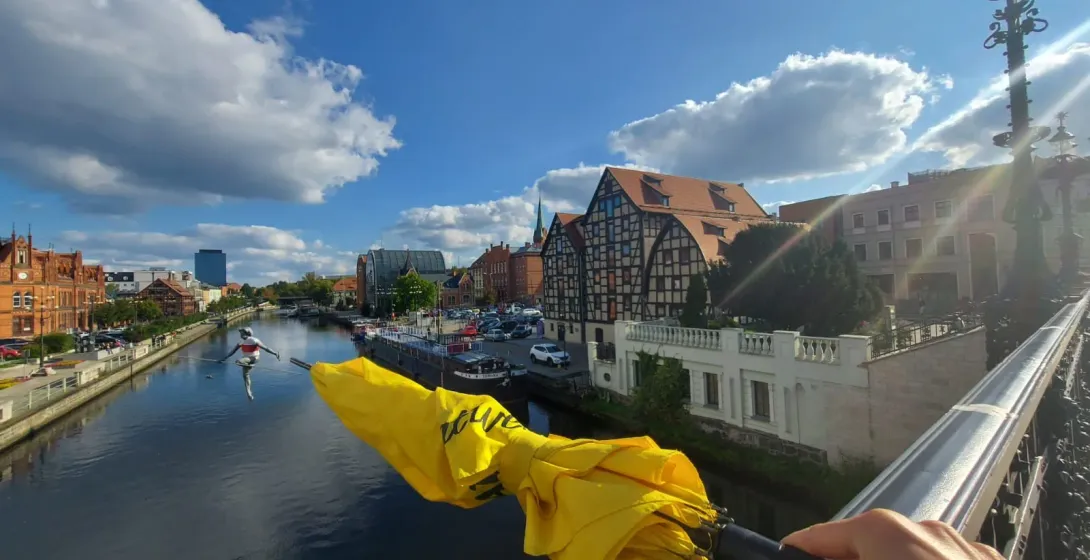 A yellow umbrella held on a bridge overlooking a river with historic buildings and a tightrope walker statue.