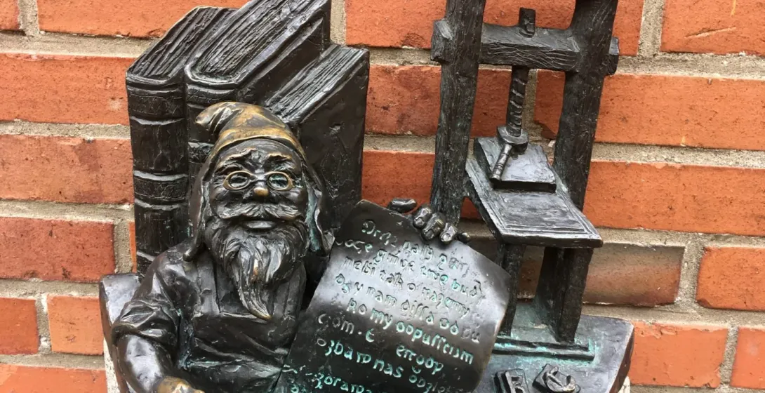 Brązowa figurka krasnala we Wrocławiu, siedząca przy prasie drukarskiej i książkach.