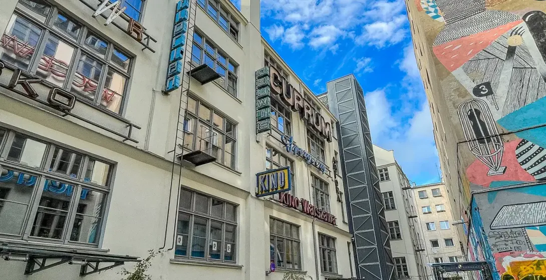 Wąska brukowana uliczka we Wrocławiu, z budynkiem z neonowymi szyldami po lewej i dużym abstrakcyjnym muralem po prawej. W...