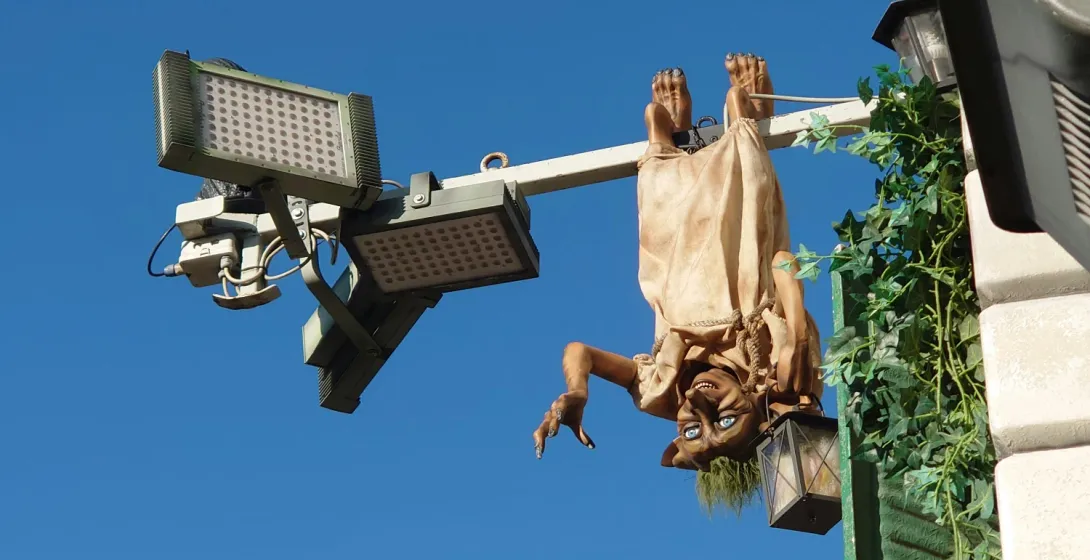 Una figura parecida a un duende cuelga boca abajo de un poste, sosteniendo un farol, con una noria al fondo.