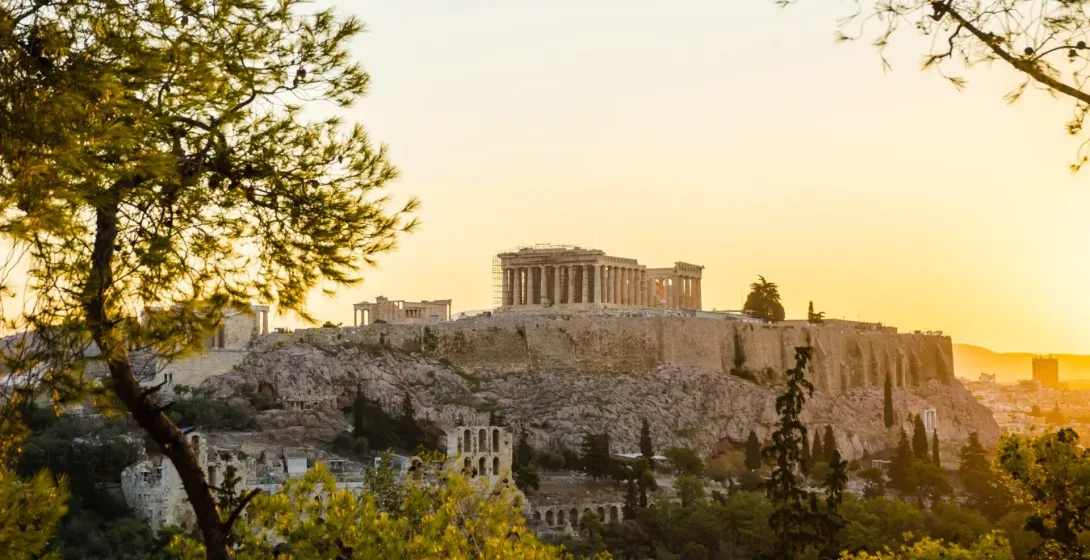 La Acrópolis de Atenas con el templo del Partenón, vista a través de los árboles contra un cielo dorado al atardecer.