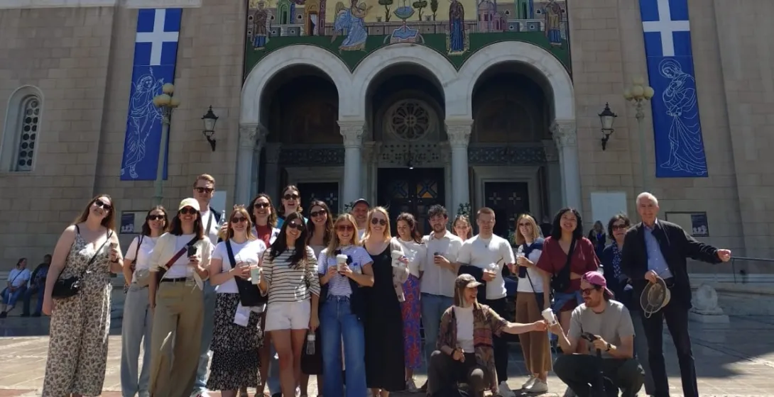 A tour group smiles for a photo in front of the Metropolitan Cathedral of Athens on a sunny day.