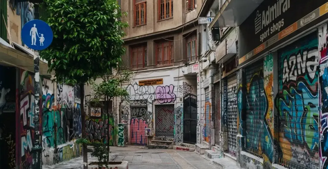 Narrow urban alley with buildings and shop shutters covered in colorful graffiti. Small trees in planters and a pedestrian...