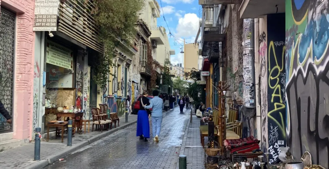 Paved street in Athens with buildings covered in graffiti and a flea market stall on the right. Two people walk away.
