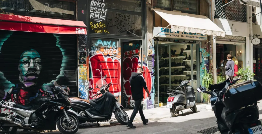 A sunny street in Athens with a large mural on a shop shutter, parked motorcycles, and a person walking past storefronts.