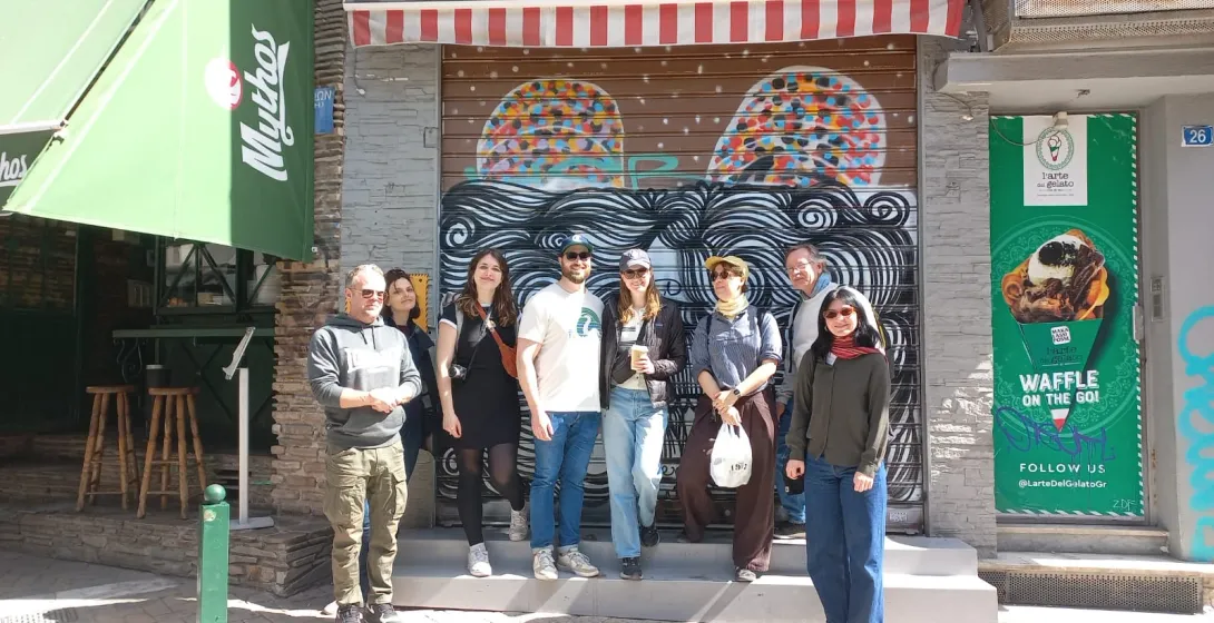 A group of tourists poses for a photo in front of a closed shop with graffiti on the streets of Athens.