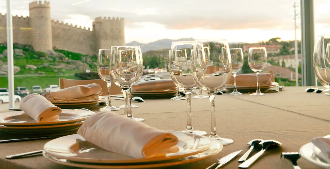Una mesa de comedor con copas y platos, con vistas a las murallas medievales de Ávila, España, al atardecer.
