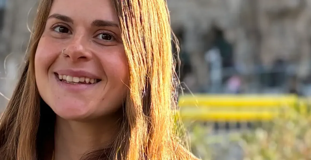 Smiling female tour guide in Barcelona with a brown coat, lanyard, and the Sagrada Familia in the blurred background.