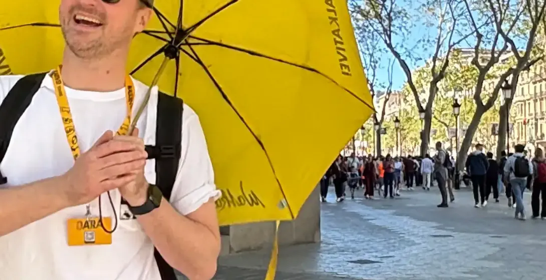 Guía turístico sonriente con paraguas amarillo de Walkative en una calle soleada de Barcelona. Edificio modernista ornamen...