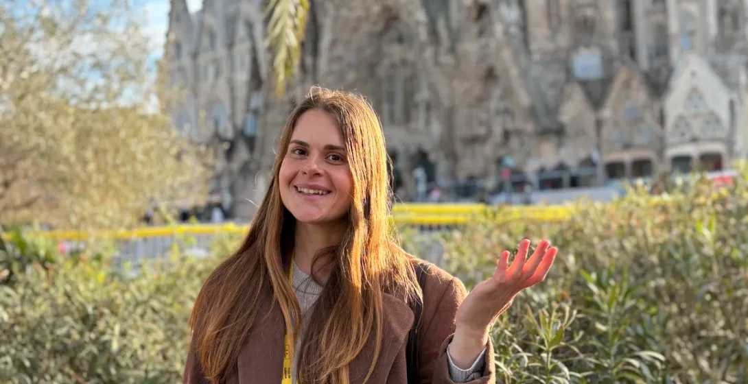 Uśmiechnięta przewodniczka Walkative w brązowym płaszczu przed bazyliką Sagrada Família Gaudiego w Barcelonie, obok palmy.
