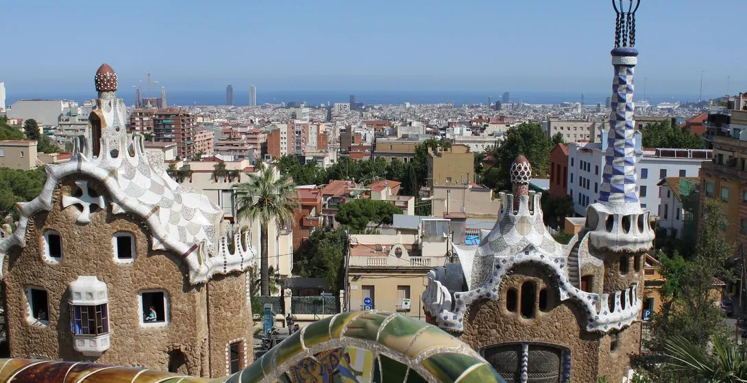 Blick vom Park Güell auf zwei Gaudi-Gebäude mit Mosaikdächern, eine bunte Fliesenmauer und Barcelonas Stadtbild mit Meer.