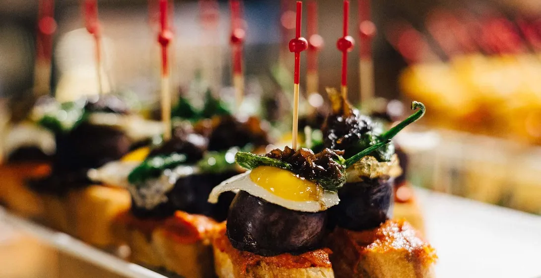 Nahaufnahme eines Tellers Pintxos, kleiner offener Sandwiches mit verschiedenen Belägen, gehalten von roten Zahnstochern. ...