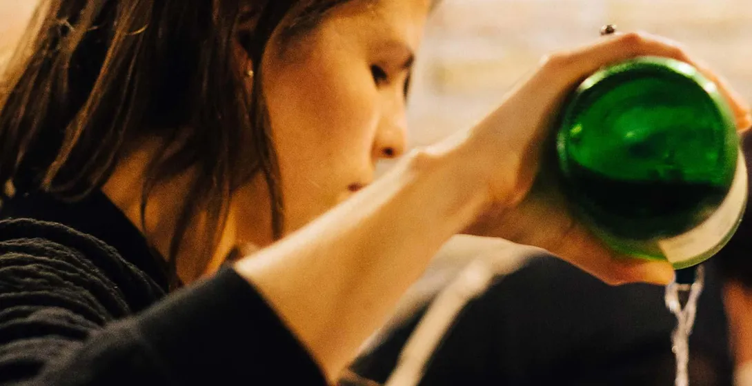 Persona sirviendo sidra de una botella verde en un vaso, creando espuma. Pared de piedra borrosa al fondo.