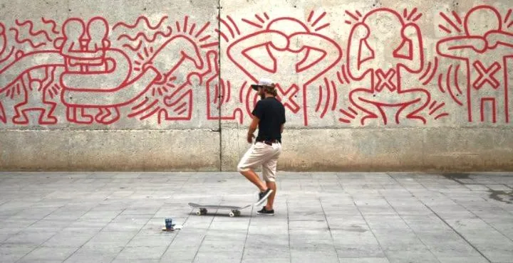 Person with skateboard in front of a concrete wall with red Keith Haring-style figures. A small blue object is on the ground.