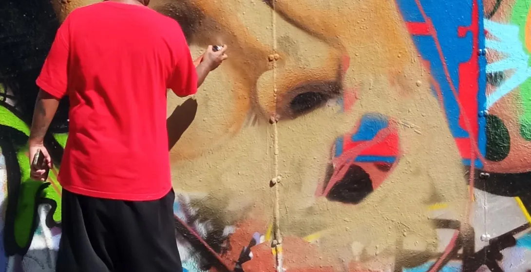 Osoba w czerwonej koszulce malująca sprayem duży mural twarzy kobiety na ścianie pokrytej graffiti. Puszki z farbą na ziemi.