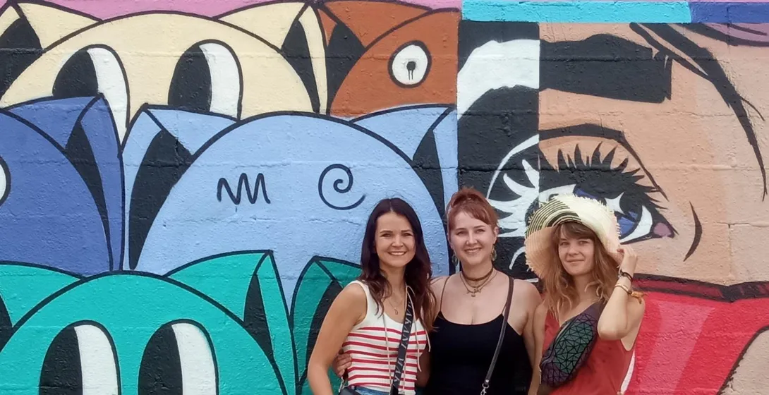 Trzy uśmiechnięte kobiety stojące przed kolorową ścianą pokrytą abstrakcyjnym graffiti i dużym muralem ze stylizowanymi twarz