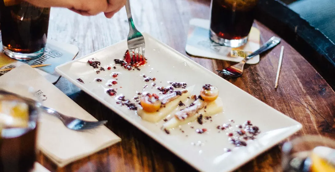 Manos de personas alrededor de una mesa de madera con platos de tapas y bebidas. Una persona come un plato con un tenedor.