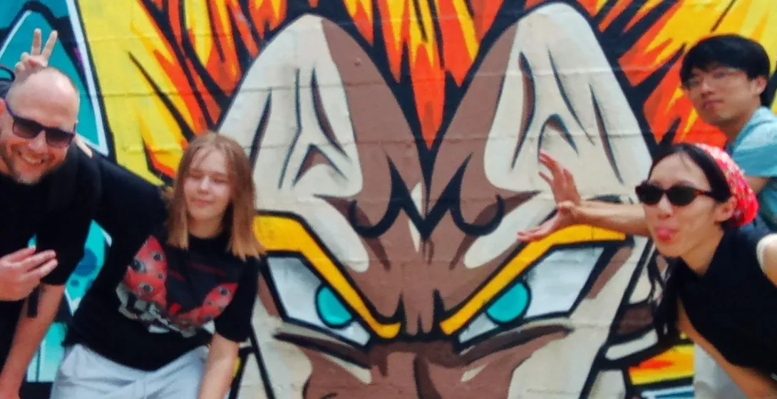 Four people posing in front of a colorful graffiti mural of a character with spiky yellow hair and blue eyes, under a clea...