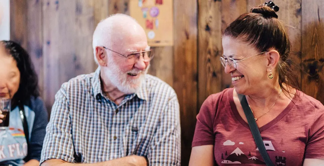 Dos viajeros sonrientes, un hombre mayor y una mujer, conversando en un ambiente informal de bar con pared de madera y cartel