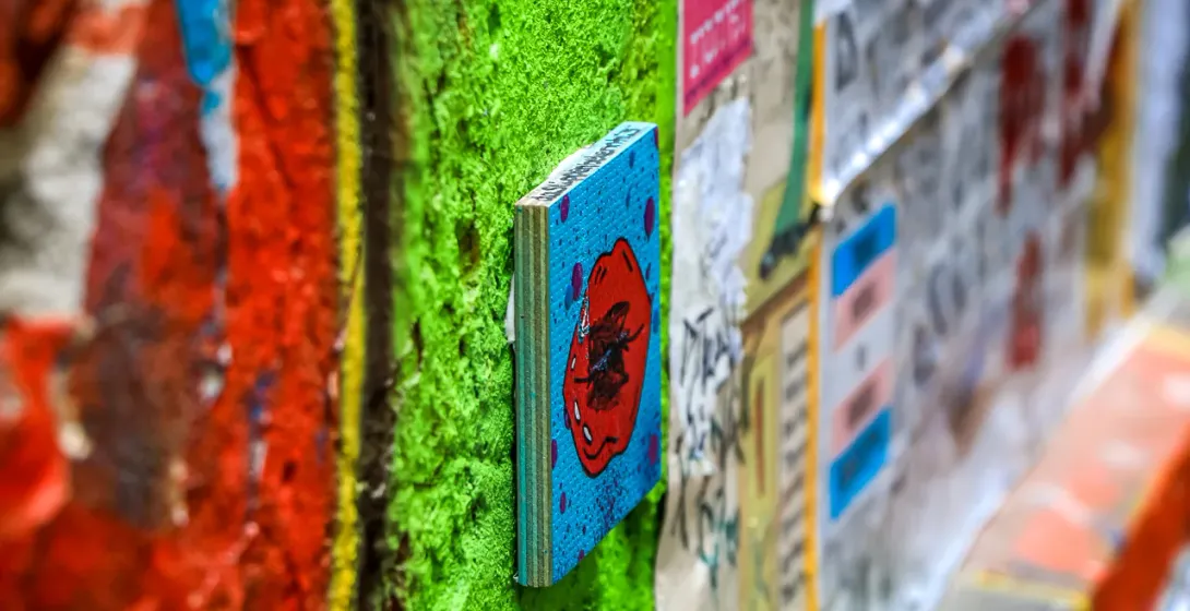 Close-up of a small wooden artwork on a bright green textured wall, surrounded by layers of paint and peeling posters.