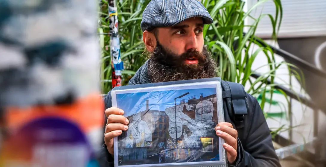 A tour guide with a beard and flat cap holds up a photograph of a large street art mural in Berlin.