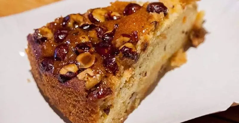 Ein Stück Kuchen mit glänzender Glasur, gerösteten Haselnüssen und getrockneten Beeren auf einem weißen Teller.