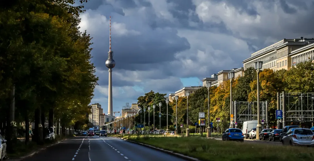 Street view of the Berlin TV Tower (Fernsehturm) on a cloudy day, with cars on the road and autumn trees.
