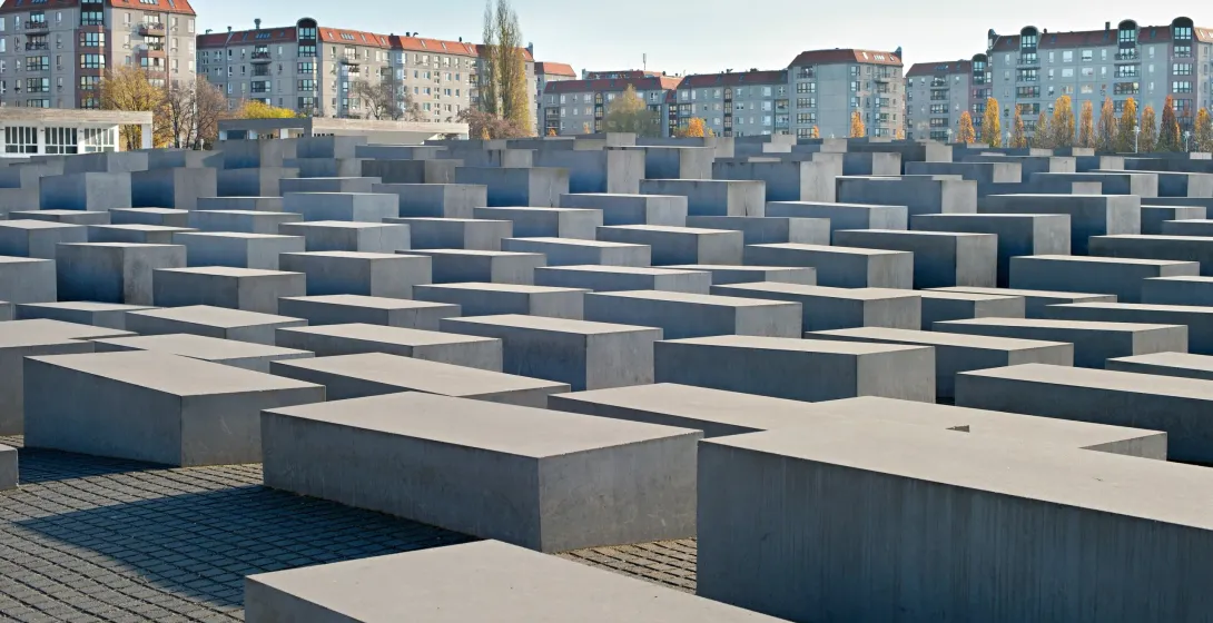 Field of concrete stelae of varying heights, forming paths, with modern apartment buildings and trees in the background.