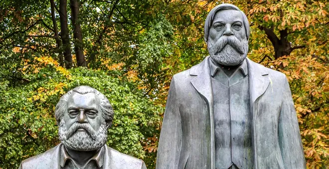 Bronze statues of Karl Marx and Friedrich Engels in Berlin, with green and autumnal trees in the background.