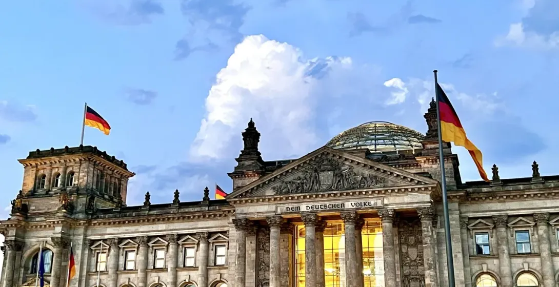 The Reichstag Building in Berlin at dusk, with German flags waving under a cloudy blue and white sky.