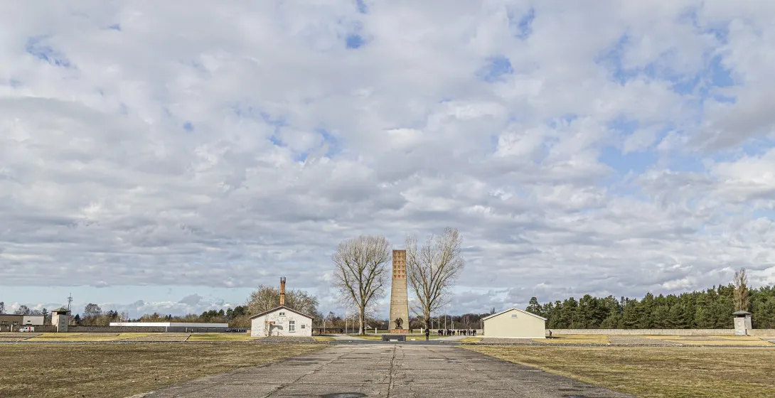 Camino de hormigón hacia un alto monumento triangular de ladrillo, flanqueado por dos árboles desnudos, con edificios y ca...