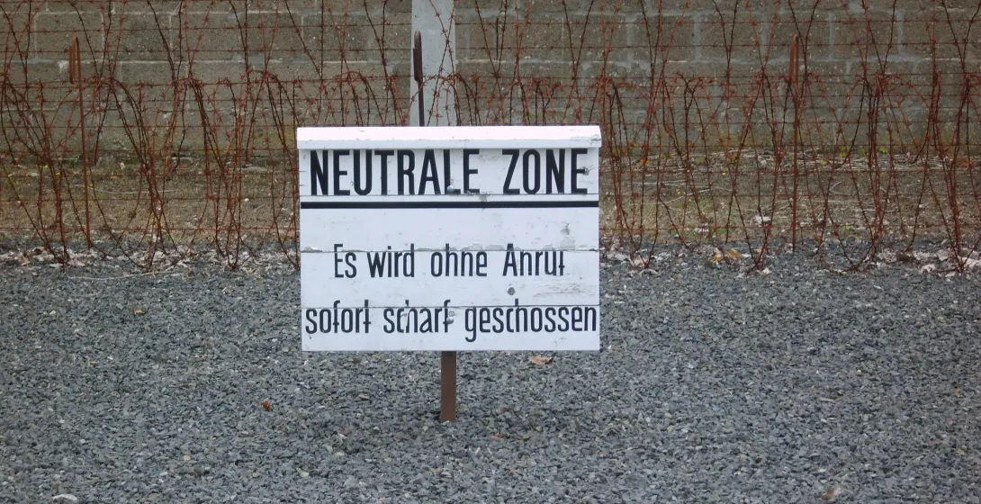 Cartel blanco con "NEUTRALE ZONE" y texto de advertencia en alemán, frente a valla de alambre de espino y muro de hormigón...