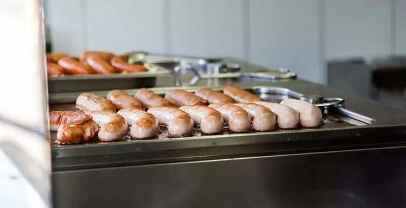 Würstchen braten auf einer Metallplatte in einer Küche mit weißen Fliesen. Eine Grillzange ist sichtbar.
