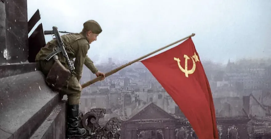 Soviet soldier planting a red flag with hammer and sickle on a destroyed building, city ruins in background.