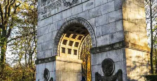 Large grey stone archway monument with carved inscriptions and reliefs, a person walks through it in a park setting.