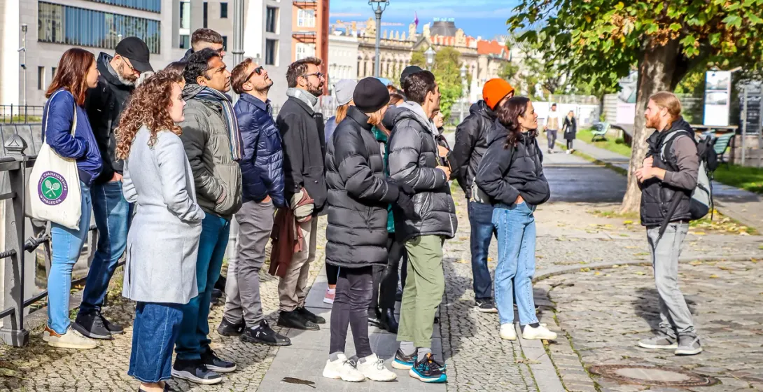 A group of tourists stands on a cobbled street listening to a tour guide, with modern and historic buildings in the backgr...