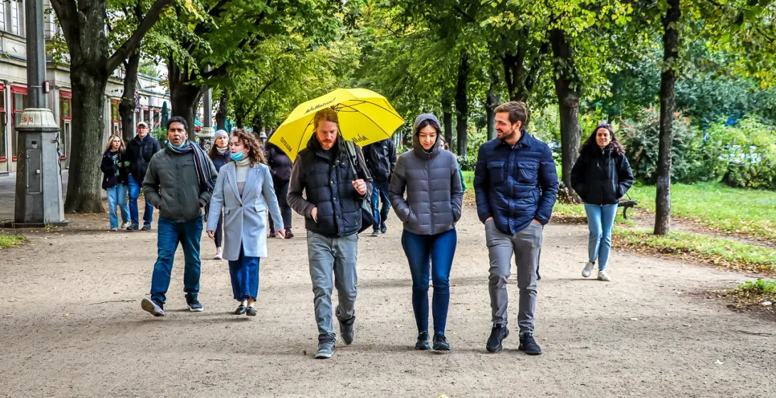 Grupo de personas caminando por un ancho sendero de tierra bordeado de árboles. Edificios a la izquierda, una persona sost...
