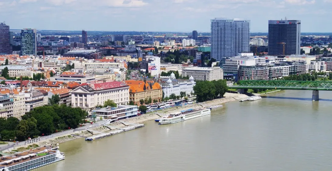 Aerial view of Bratislava cityscape along the Danube River, featuring modern skyscrapers, historic buildings, and a green arc